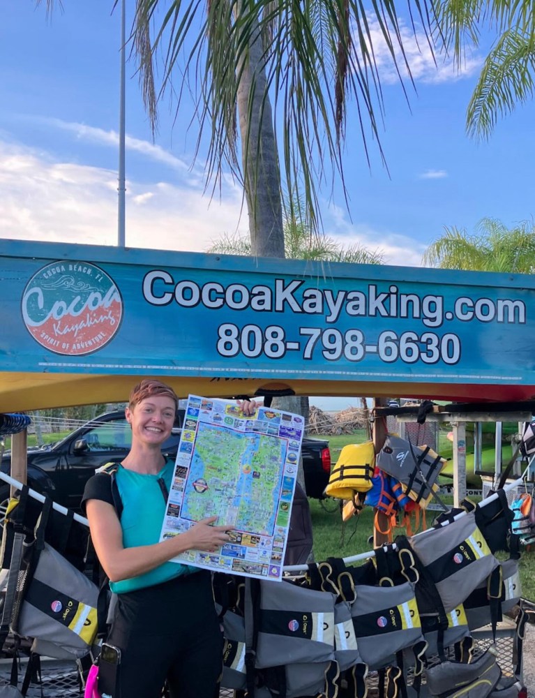 Person holding a map, standing by a sign reading 'CocoaKayaking.com' with life jackets hanging nearby.