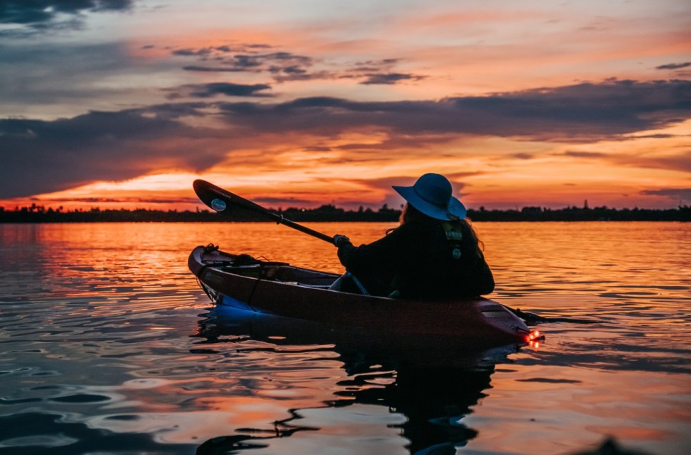 Silhouette of a person kayaking on a lake at sunset with a dramatic orange sky.