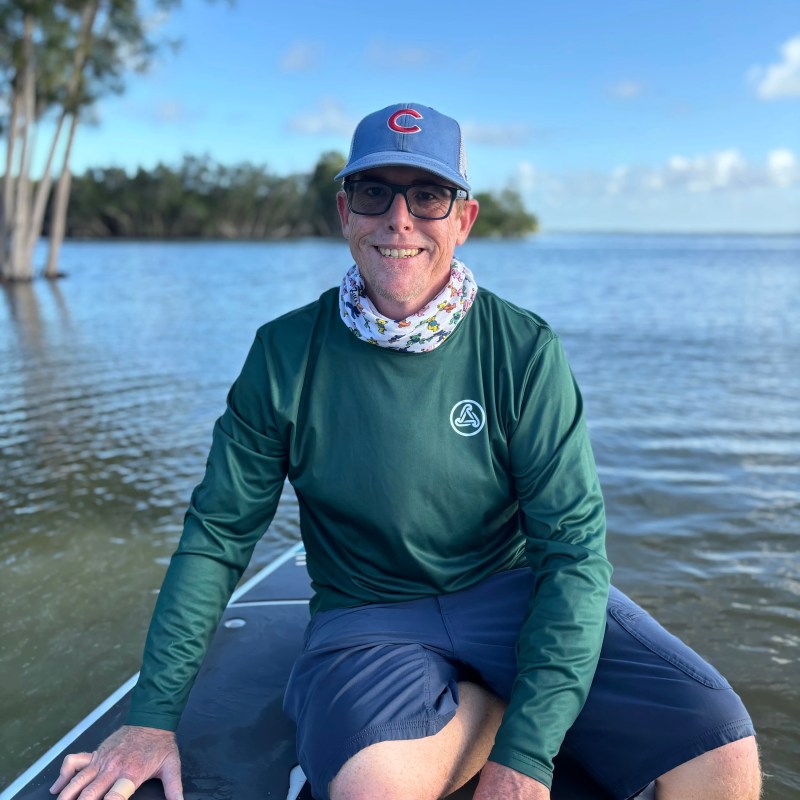 Jack Man in green shirt and blue cap kneeling on a boat with a lake and trees in the background.