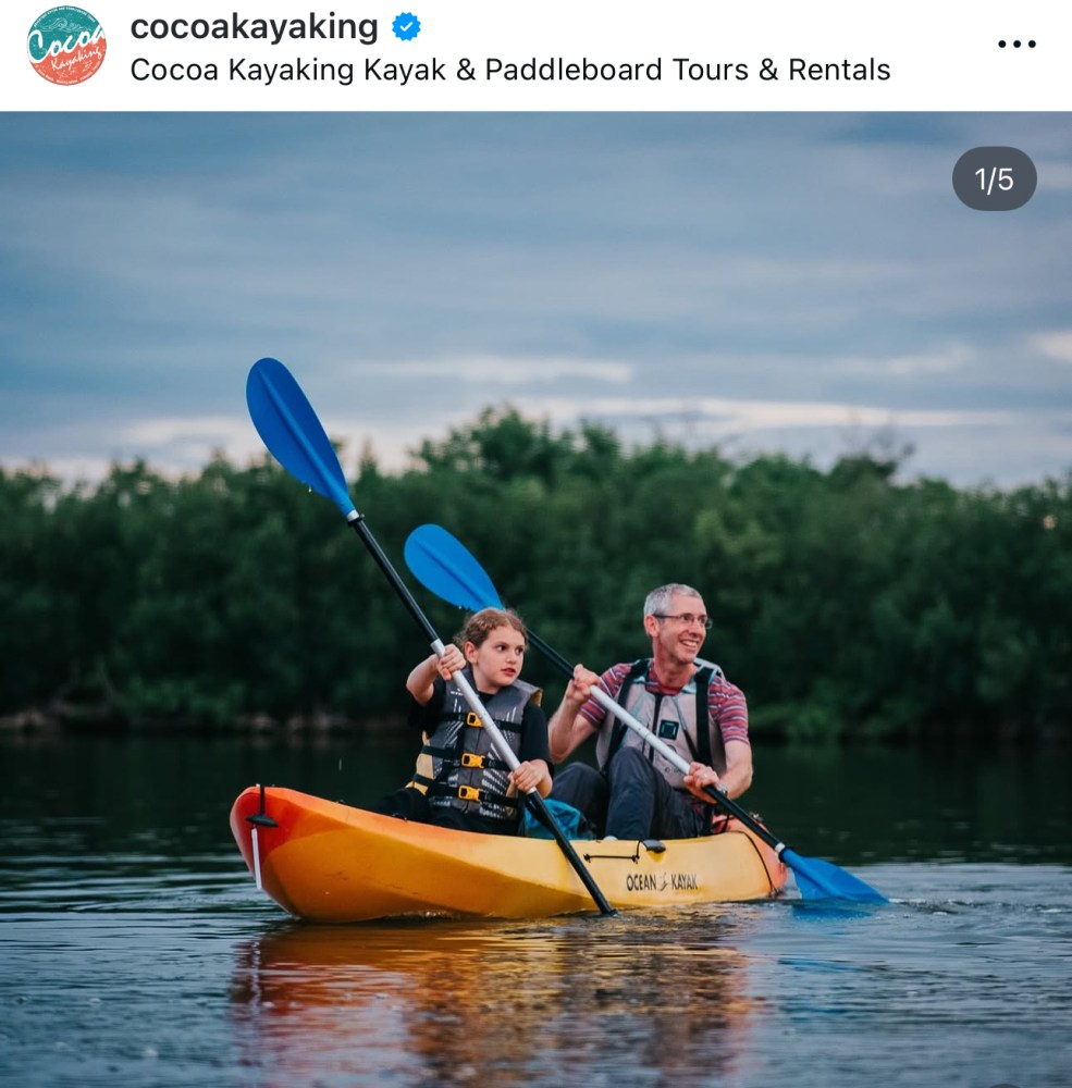 Two people kayaking on a calm river with trees in the background.