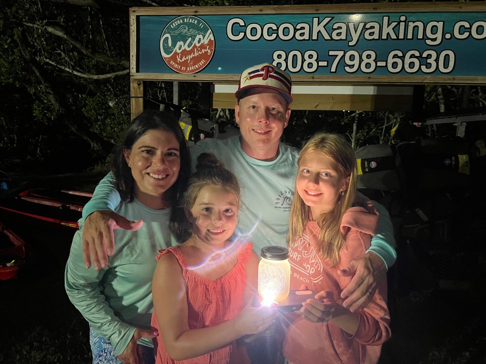 Family of four holding a glowing jar, standing under a Cocoa Kayaking sign at night.