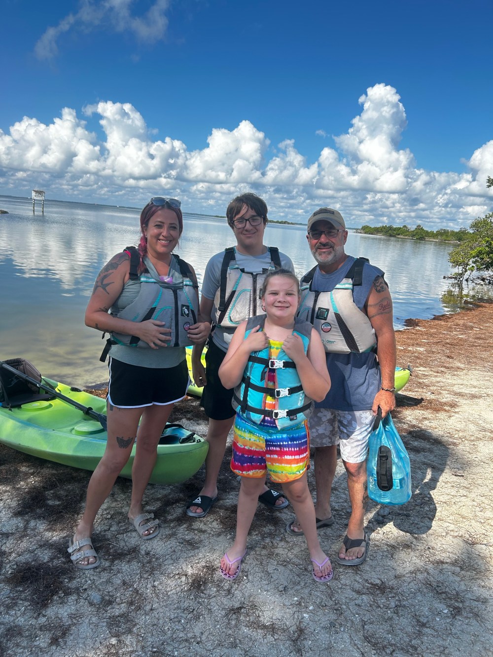 Four people in life jackets stand near kayaks by a lake under a blue sky with clouds.