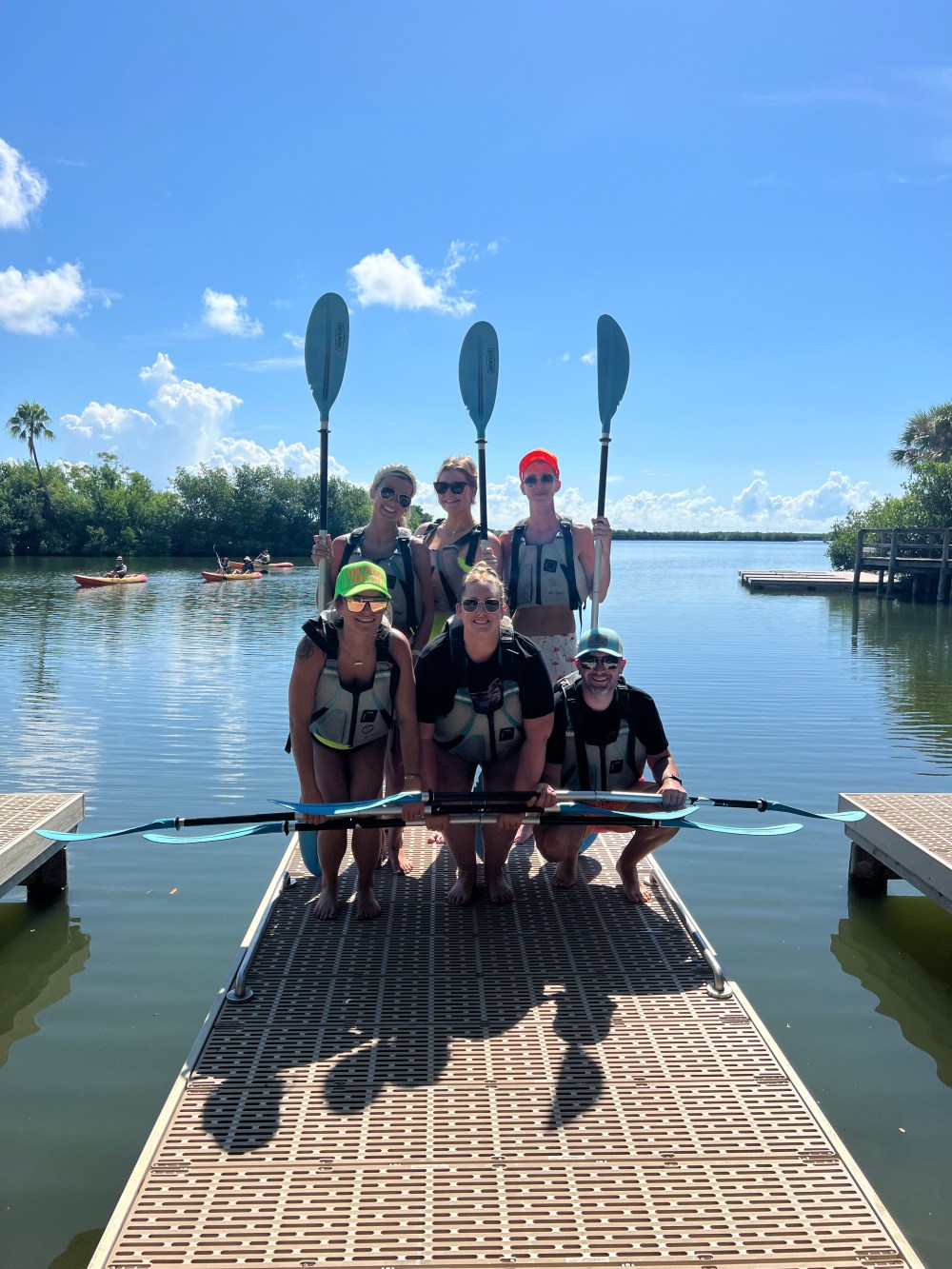 Group of six people holding paddles on a dock with water and kayaks in the background.