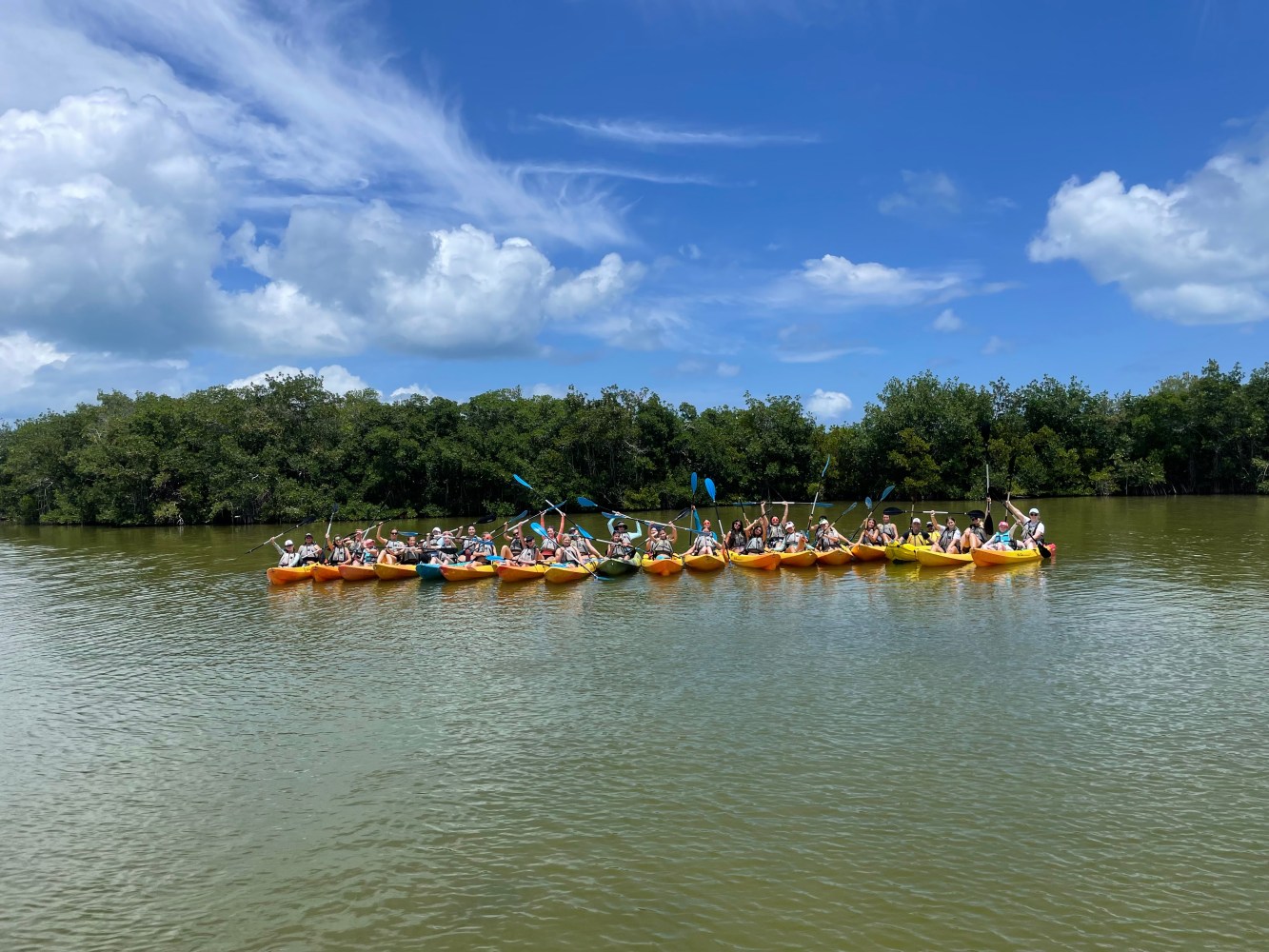 Group of kayakers on a river with trees in the background under a blue sky.