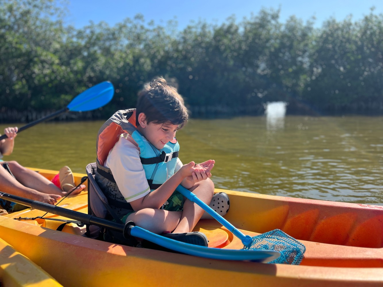 Child in life jacket sitting in kayak on water, holding a small object.