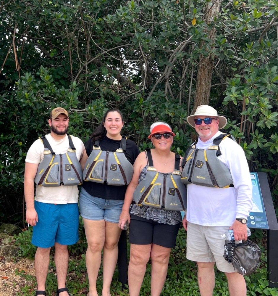 Four people wearing life jackets stand in front of greenery.