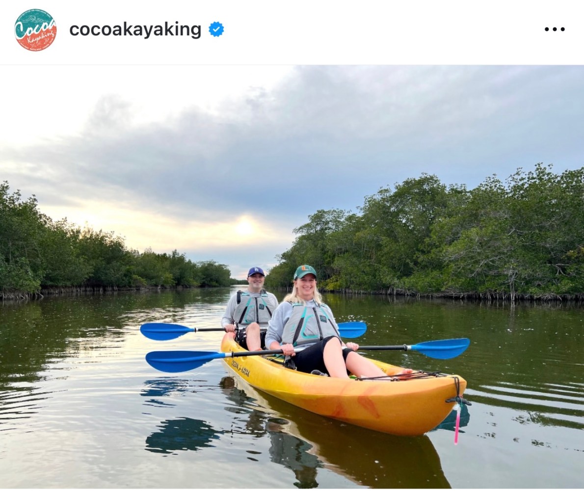 Two people kayaking on a calm waterway surrounded by lush greenery.
