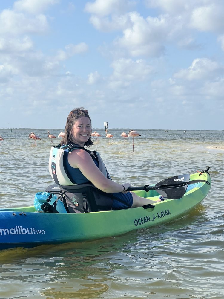 Woman smiles on green kayak in water with flamingos in the background under a cloudy sky.