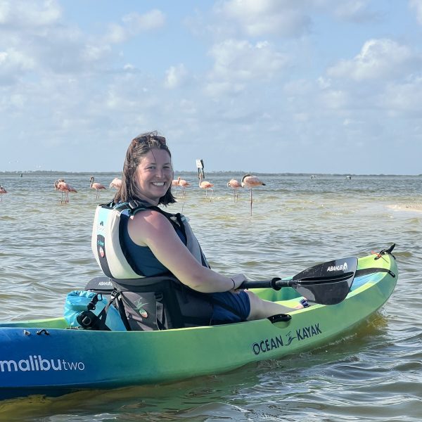 Woman smiles on green kayak in water with flamingos in the background under a cloudy sky.