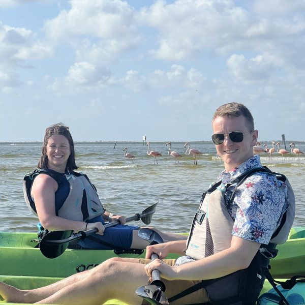 Two people kayaking with flamingos in the background under a cloudy blue sky.