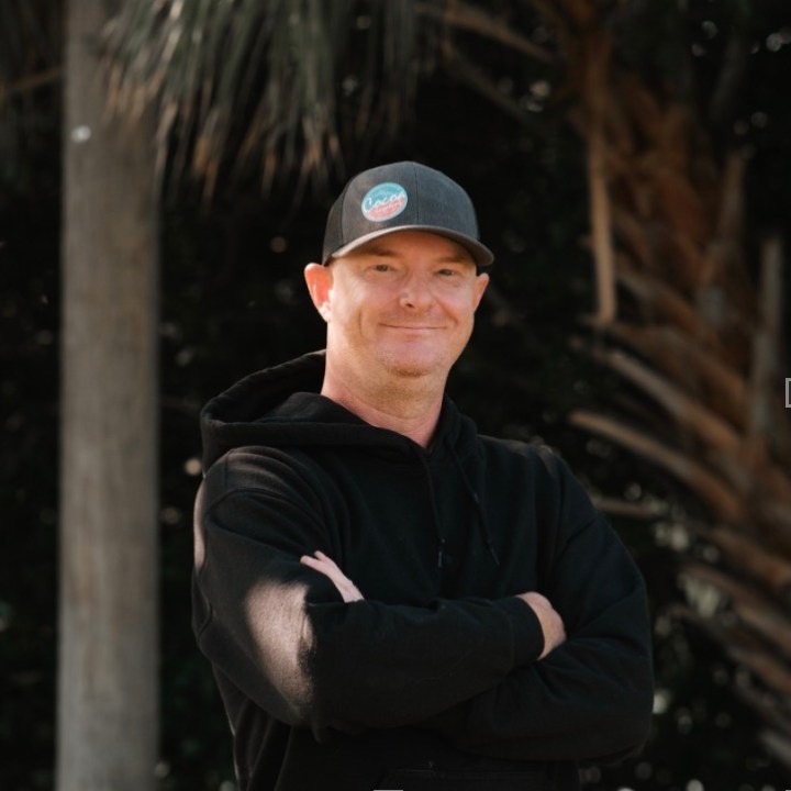 Man in hoodie and cap smiling outdoors with arms crossed.