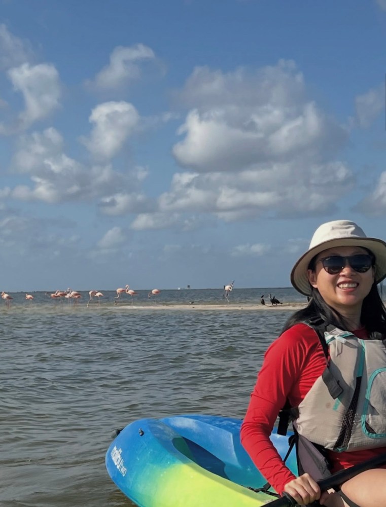 Person in kayak on water, wearing hat, with flamingos on sandbar under cloudy sky.