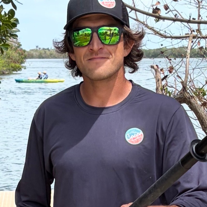 Man in sunglasses and cap holding paddle by river with kayakers in background.