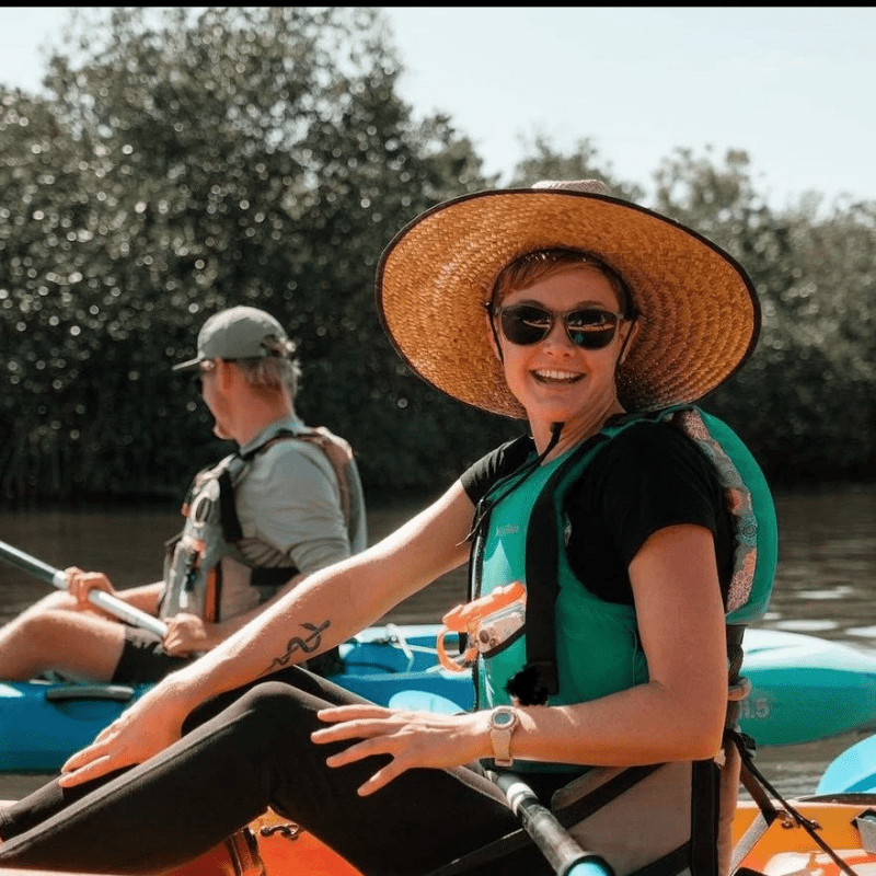 Two people kayaking on a sunny day, one wearing a wide-brimmed hat and sunglasses.