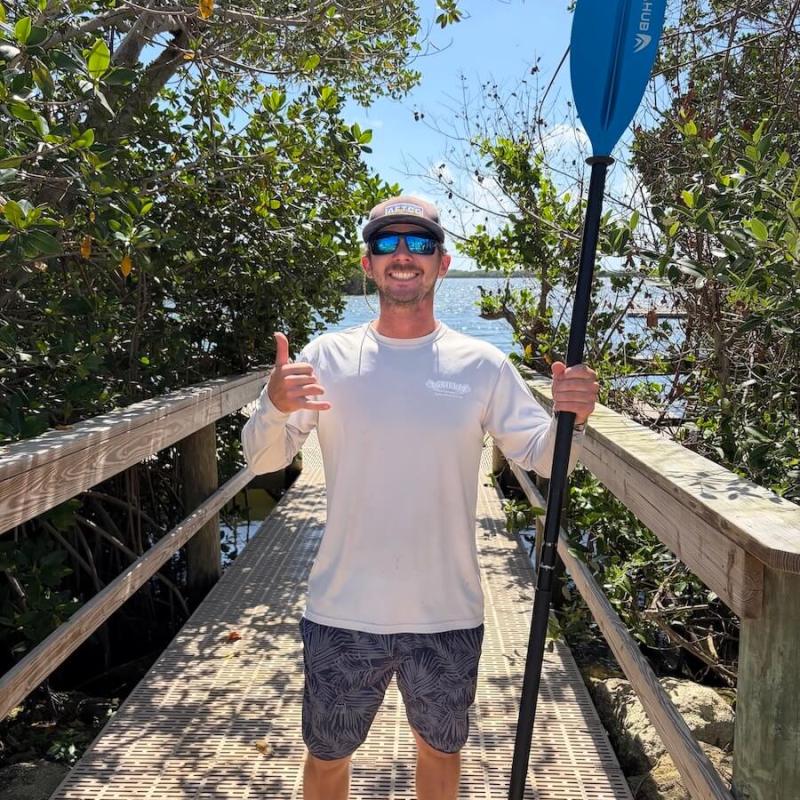Man on boardwalk holding a paddle, wearing sunglasses and a hat, trees and water in the background.