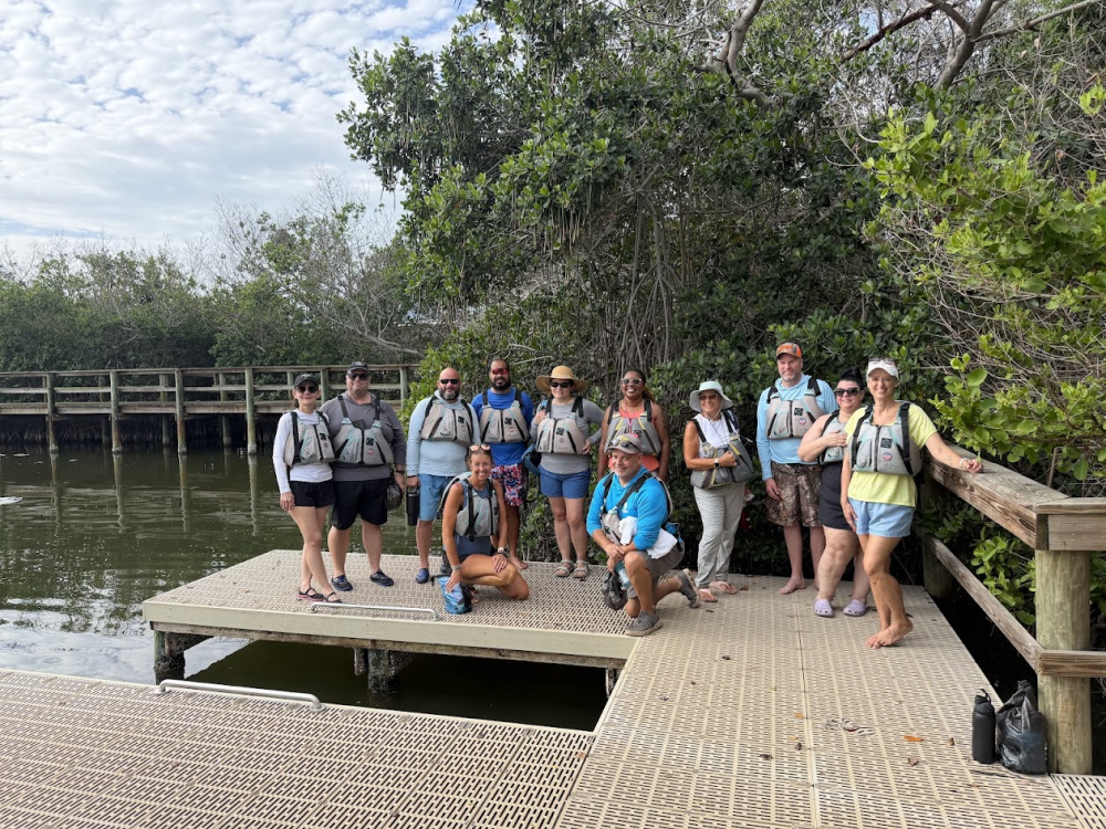 Group of people posing on a wooden dock by water and trees.
