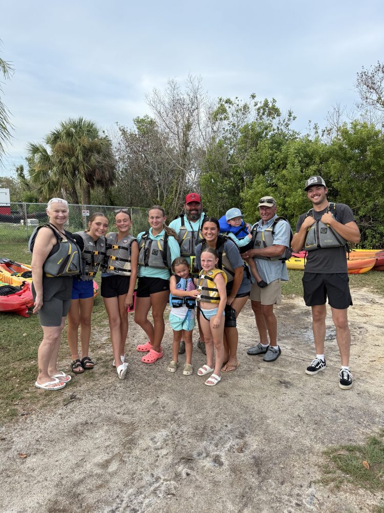 Group of people in life vests standing outdoors near kayaks and trees.