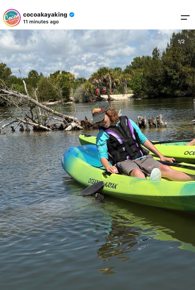 Person in a green kayak on calm water, surrounded by trees and cloudy sky.