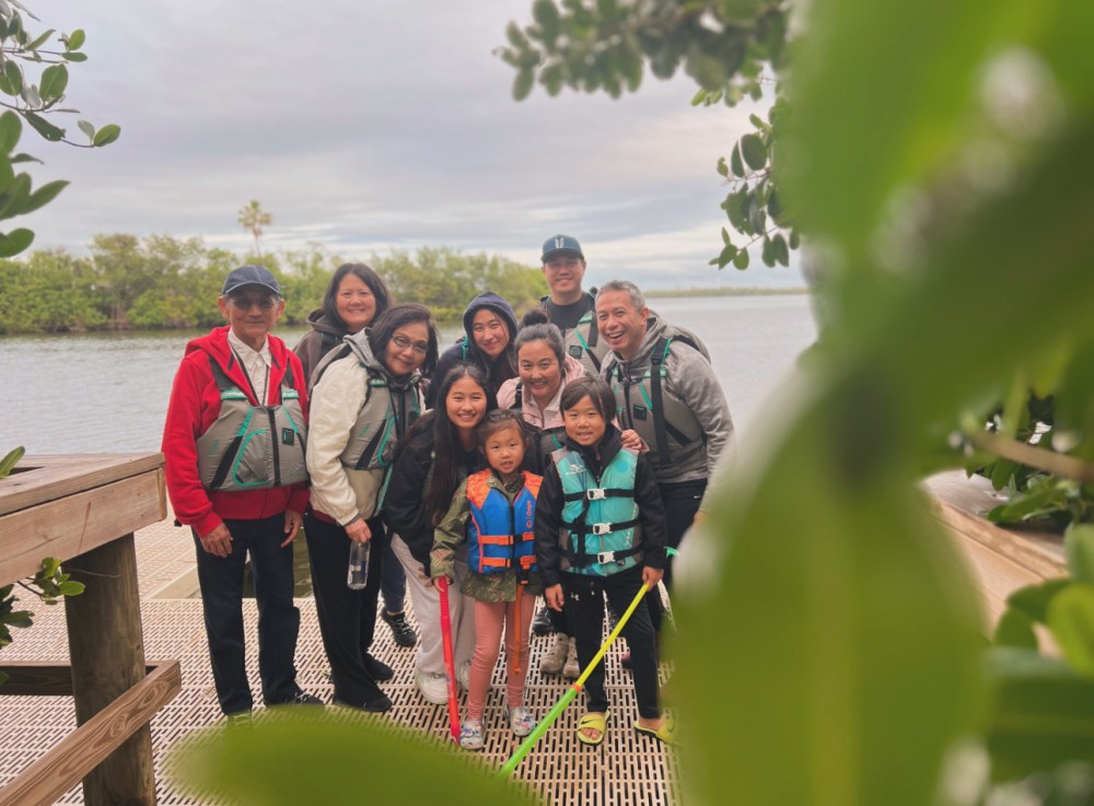 Group of people posing on a riverside walkway with life jackets and trees in the foreground.