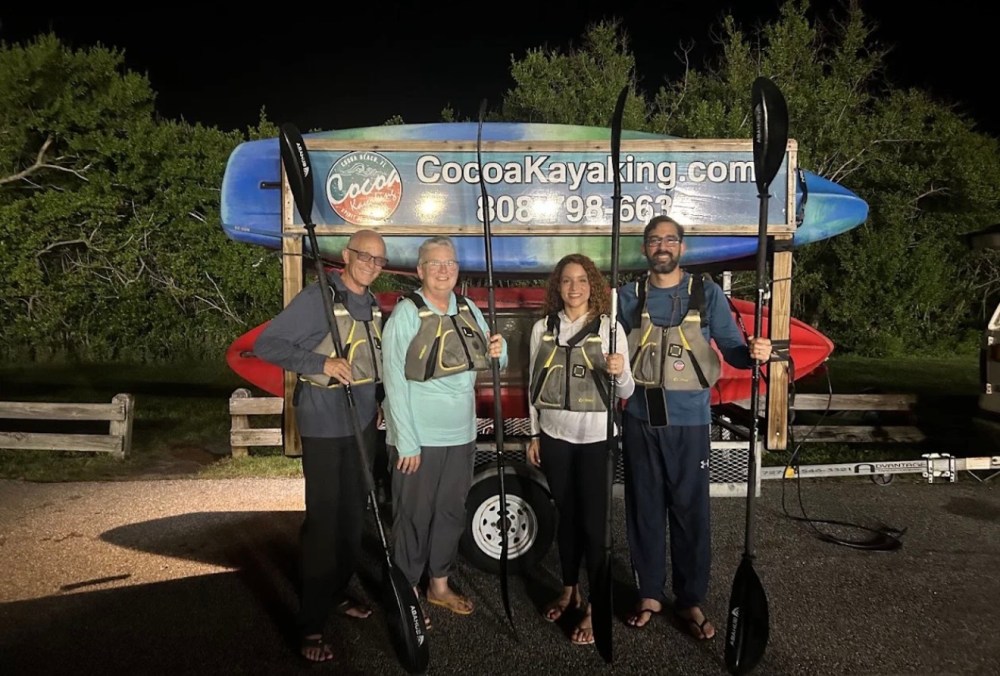 Four people in life vests holding paddles, standing by kayaks at night.