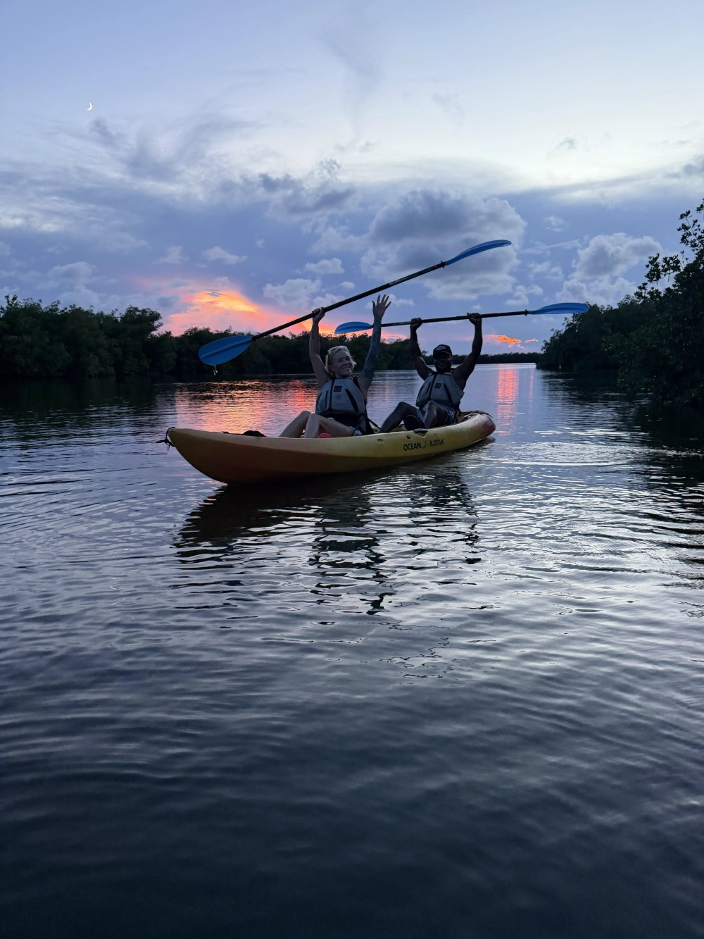 Two people kayaking on a calm lake at sunset with raised paddles.