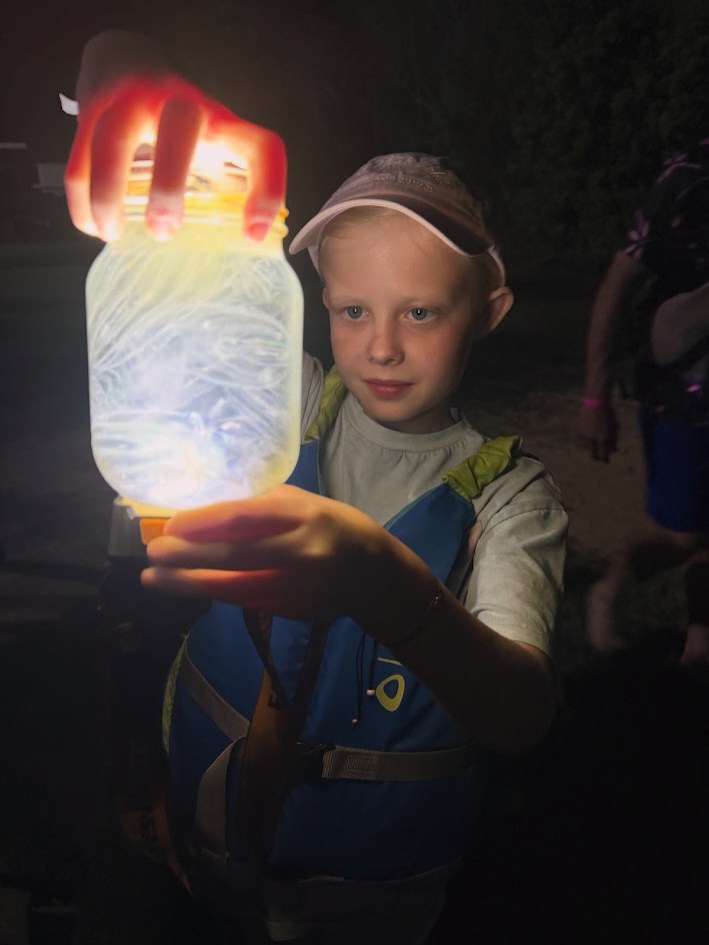 Child holding a glowing jar at night, wearing a cap and life jacket.
