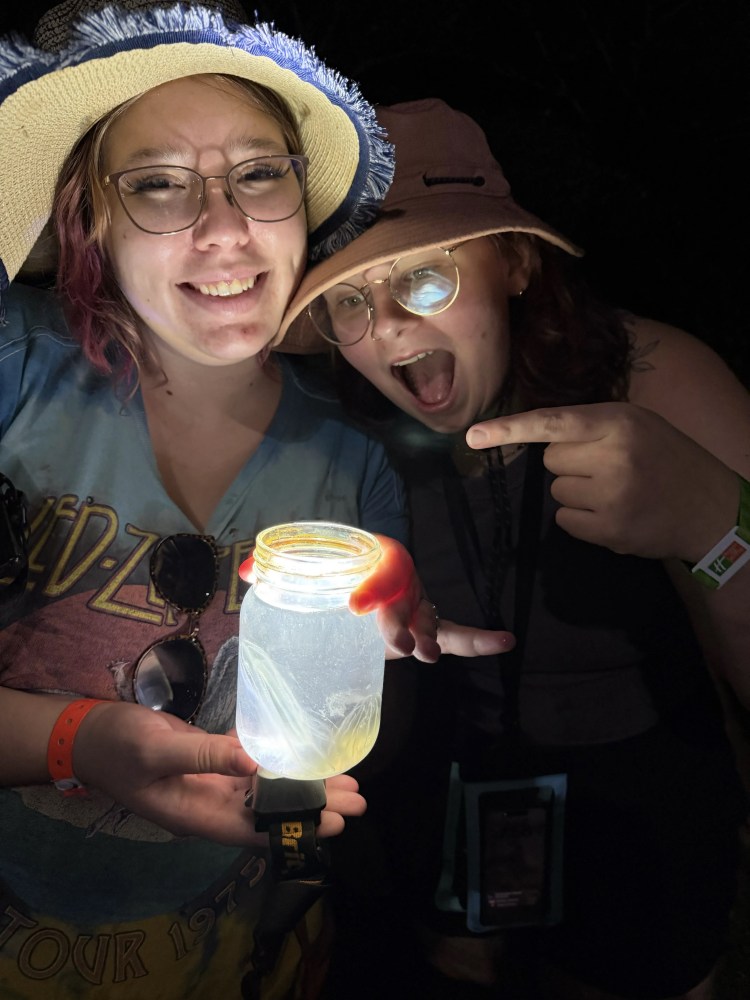 Two people with hats smiling and looking at a glowing jar in the dark.